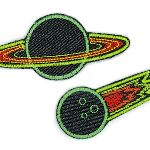 Può includere: Due toppe ricamate da stirare con disegni a tema spaziale. La prima toppa è un Saturno verde e giallo con un anello rosso. La seconda toppa è una cometa verde e gialla con una coda rossa.