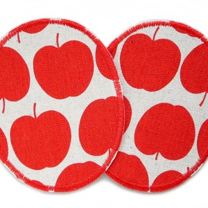 May include: Two round, white fabric coasters with red apple designs. The coasters have a stitched edge.