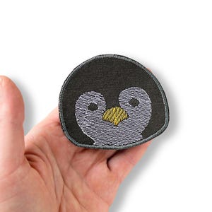 May include: Embroidered penguin face patch. The patch has a dark grey background with a stitched outline. The penguin's face is detailed with white thread for the eyes and a yellow beak. The patch is held in a hand.
