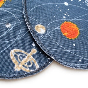 K&ouml;nnte beinhalten: Zwei runde Stoffpatches mit dunkelblauem Hintergrund und wei&szlig;en Umrissen von Planeten und Sternen. Ein Patch zeigt eine gelbe Sonne und einen roten Planeten. Der andere Patch zeigt ein gelbes Raumschiff.