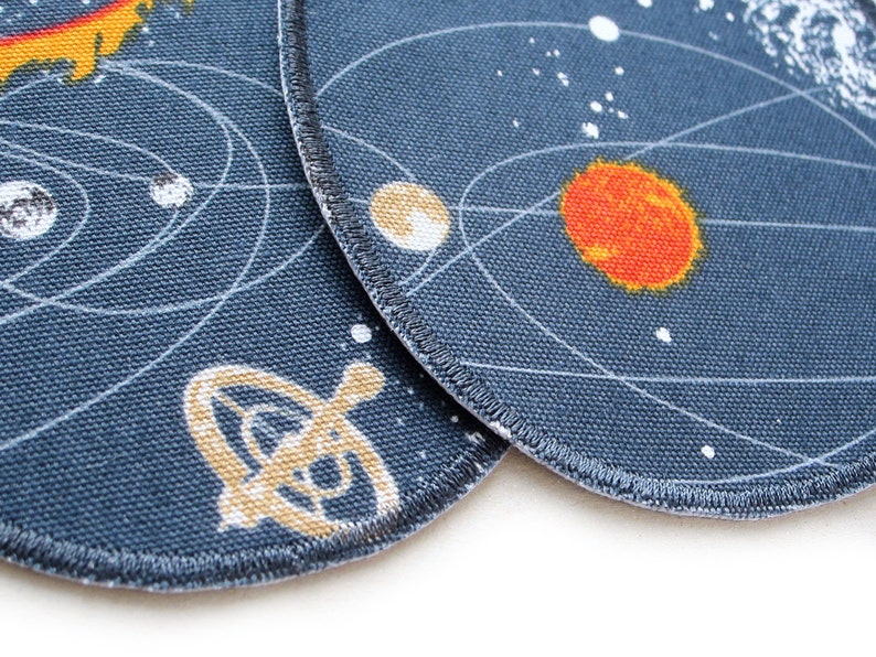 K&ouml;nnte beinhalten: Zwei runde Stoffpatches mit dunkelblauem Hintergrund und wei&szlig;en Umrissen von Planeten und Sternen. Ein Patch zeigt eine gelbe Sonne und einen roten Planeten. Der andere Patch zeigt ein gelbes Raumschiff.