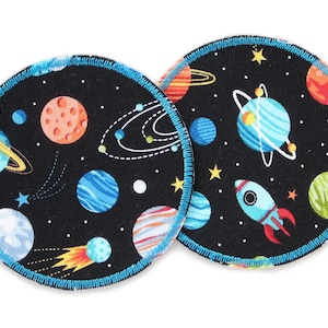 Könnte beinhalten: Zwei runde, schwarze Still-Einlagen aus Stoff mit einem Weltraum-Motiv. Der Druck zeigt Planeten, Sterne, eine Rakete und Kometen. Die Einlagen haben einen blauen Rand.
