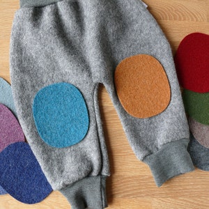 May include: Grey wool trousers featuring a ribbed waistband and cuffs, with a blue felt patch on the left knee and an orange felt patch on the right knee. Several additional felt patches in various colours are arranged around the trousers.