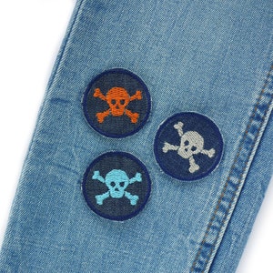 May include: Three embroidered skull and crossbones patches on a blue denim background. The patches are in different colours: orange, grey, and blue.
