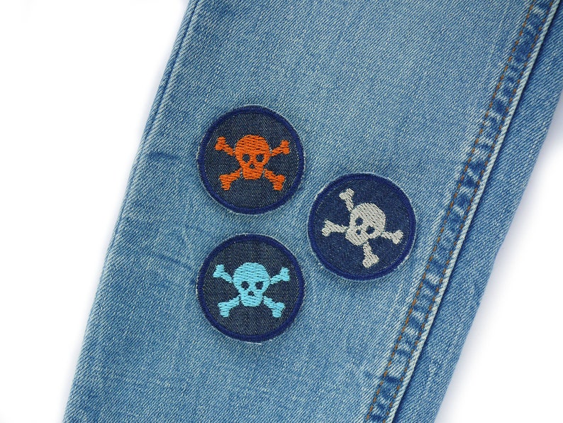 May include: Three embroidered skull and crossbones patches on a blue denim background. The patches are in different colours: orange, grey, and blue.