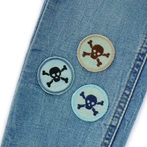 May include: Three embroidered patches featuring a skull and crossbones design, on a blue denim background. The patches are in different colours: light blue, brown, and dark blue.