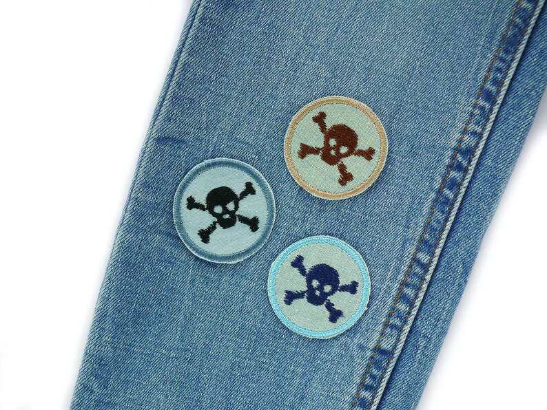 May include: Three embroidered patches featuring a skull and crossbones design, on a blue denim background. The patches are in different colours: light blue, brown, and dark blue.