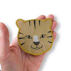 Op de afbeelding: Geborduurde tijgerkop patch met een bruinachtige achtergrond, zwarte strepen en een witte neus. De patch heeft een gele rand en wordt in een hand gehouden. Perfect voor kleding of accessoires.