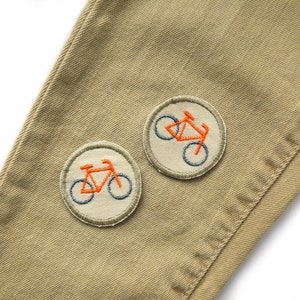 May include: Two embroidered patches on a beige fabric background. Each patch features a bicycle with orange and blue stitching.