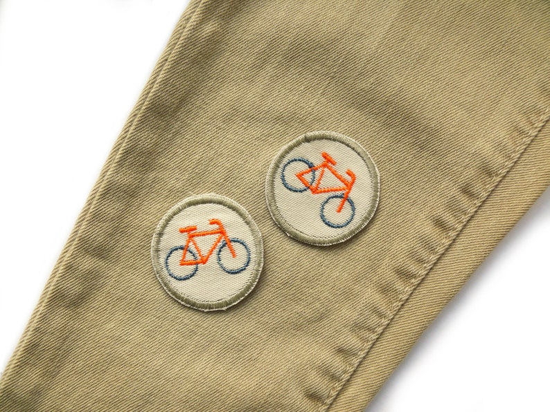 May include: Two embroidered patches on a beige fabric background. Each patch features a bicycle with orange and blue stitching.