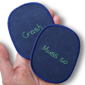 Pode incluir: Dois emblemas de tecido azul-marinho em formato oval com bordas costuradas em azul escuro. Um emblema apresenta a palavra "Crash" bordada em linha verde brilhante, enquanto o outro apresenta as palavras "Muss so" bordadas em linha verde.
