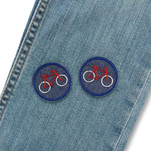 May include: Close-up of light blue denim jeans with two circular patches. Each patch features a red bicycle outline with white wheels, set against a dark blue background. The jeans have visible stitching.