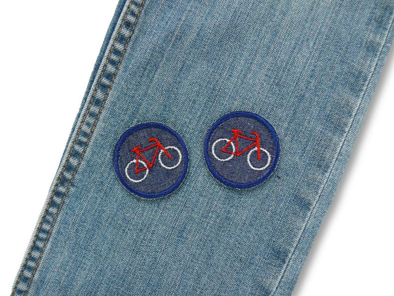 May include: Close-up of light blue denim jeans with two circular patches. Each patch features a red bicycle outline with white wheels, set against a dark blue background. The jeans have visible stitching.