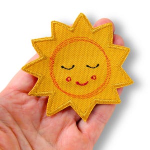Könnte beinhalten: Ein gelber, gestickter Sonnenfleck mit einem lächelnden Gesicht. Die Sonne hat ein kreisförmiges oranges Gesicht mit geschlossenen Augen, rosigen Wangen und einem lächelnden Mund. Die Sonnenstrahlen sind spitz und der Aufnäher wird in einer Hand gehalten.
