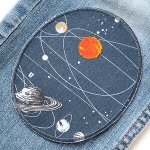 K&ouml;nnte beinhalten: Ein blauer Jeansstoff mit einem runden Patch, der ein stilisiertes Sonnensystem mit einer gro&szlig;en orangefarbenen Sonne, einem schwarz-wei&szlig;en Planeten mit Ringen und wei&szlig;en Sternen auf dunkelblauem Hintergrund darstellt.