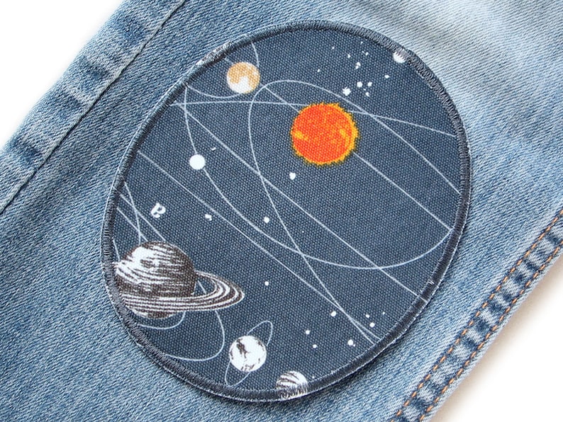 K&ouml;nnte beinhalten: Ein blauer Jeansstoff mit einem runden Patch, der ein stilisiertes Sonnensystem mit einer gro&szlig;en orangefarbenen Sonne, einem schwarz-wei&szlig;en Planeten mit Ringen und wei&szlig;en Sternen auf dunkelblauem Hintergrund darstellt.