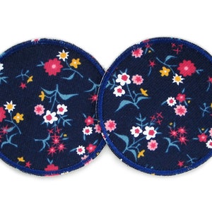 Pode incluir: Dois discos de amamentação redondos de tecido azul marinho com estampa floral em rosa, branco e amarelo. Os discos têm uma borda azul escura.