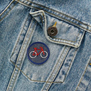 May include: Close-up of a light blue denim jacket with a small, round patch featuring a red bicycle design on a navy blue background. The jacket has a buttoned pocket and a metal button.