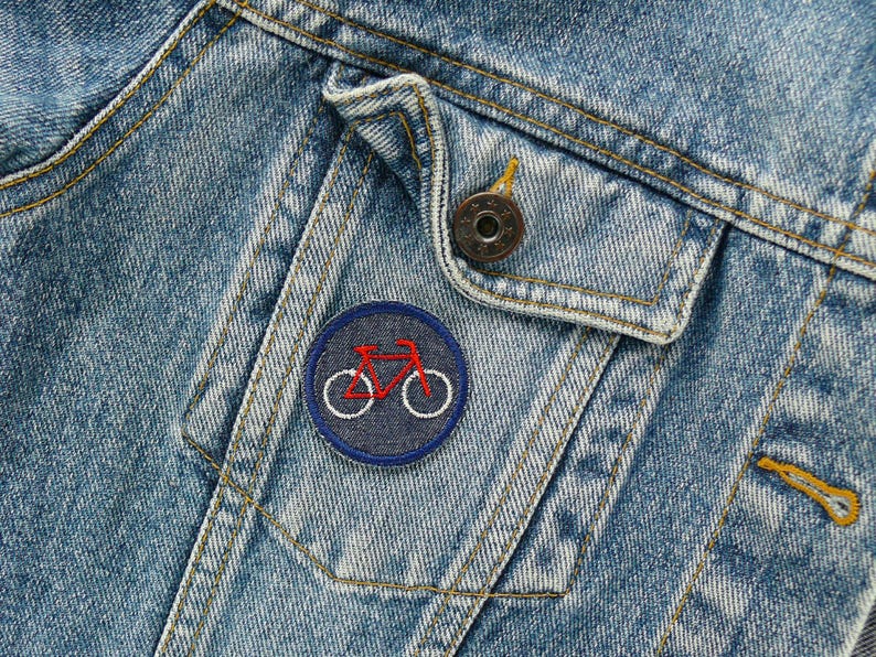 May include: Close-up of a light blue denim jacket with a small, round patch featuring a red bicycle design on a navy blue background. The jacket has a buttoned pocket and a metal button.