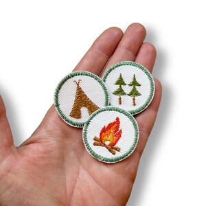 May include: Three circular embroidered patches with white backgrounds and green stitched borders. One patch features a brown teepee, another shows two green pine trees, and the third displays a bright orange and yellow campfire with brown logs.