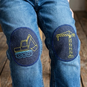 Könnte beinhalten: Blaue Jeans mit zwei dunkelblauen Aufnähern, die mit Baumaschinen bestickt sind. Ein Aufnäher zeigt einen gelben Bagger, der andere einen gelben Kran. Die Aufnäher sind kreisförmig und auf die Knie genäht.