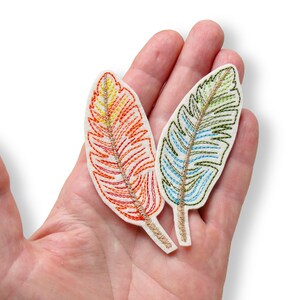May include: Two embroidered feather patches on a white fabric base. One patch features a gradient of yellow, orange, and red stitching, while the other displays green and blue thread. Both have a central gold-coloured embroidered spine detail.