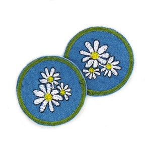 Könnte beinhalten: Zwei blaue Jeans-Patches mit grüner Naht, jeder mit einer weißen gestickten Gänseblümchen-Gruppe mit gelben Zentren.