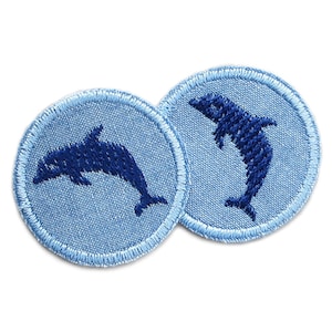 Peut inclure: Deux écussons ronds brodés bleu clair avec un motif de dauphin bleu foncé. Les dauphins sautent et nagent. Les écussons ont une bordure bleu clair et sont sur fond blanc.