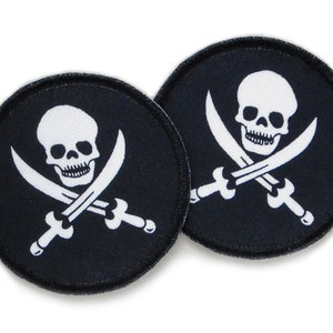 Könnte beinhalten: Zwei schwarze runde Stoffpatches mit weißen gestickten Totenkopf- und Knochenkreuz-Designs. Der Schädel hat ein zahniges Grinsen und die Knochenkreuze sind Schwerter.
