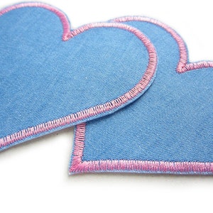 Pu&ograve; includere: Due toppe di tessuto blu a forma di cuore con bordi ricamati rosa. Le toppe si sovrappongono leggermente, evidenziando le cuciture dettagliate e il contrasto tra il tessuto blu e il filo rosa. I cuori sono su sfondo bianco.