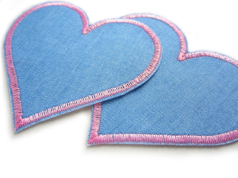 Pu&ograve; includere: Due toppe di tessuto blu a forma di cuore con bordi ricamati rosa. Le toppe si sovrappongono leggermente, evidenziando le cuciture dettagliate e il contrasto tra il tessuto blu e il filo rosa. I cuori sono su sfondo bianco.