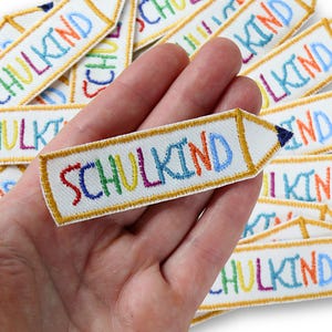 Peut inclure: Une collection d'écussons brodés blancs en forme de crayons avec des bordures jaunes et des pointes bleu foncé. Chaque écusson présente le mot SCHULKIND brodé en lettres multicolores avec du fil rouge, vert, jaune, rose et bleu.