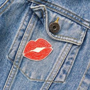 Könnte beinhalten: Jeansjacke mit rotem, gesticktem Lippen-Patch auf der Tasche. Der Patch ist detailliert gestaltet mit weißen Nähten. Die Jacke hat einen Knopfverschluss und ein klassisches Taschen-Design.