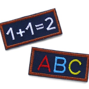 Könnte beinhalten: Zwei gestickte Aufnäher. Einer ist marineblau mit braunem Rand und sagt "1+1=2" in weißem Garn. Der andere ist marineblau mit braunem Rand und sagt "ABC" in rotem, blauem und gelbem Garn.
