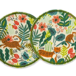 Può includere: Due sottobicchieri rotondi in tessuto con un design a tema giungla. I sottobicchieri presentano uno sfondo color crema con illustrazioni di tigri, leopardi, scimmie, fiori e fogliame. I bordi sono rifiniti con cuciture verdi.