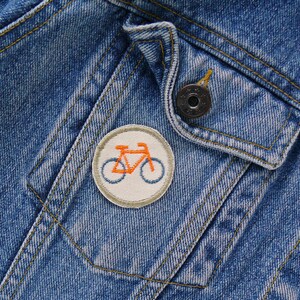 May include: A close-up of a denim jacket with a small, round, white patch featuring an orange bicycle. The patch is sewn onto the jacket's pocket flap.