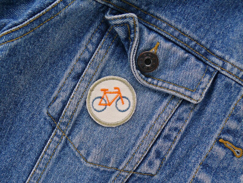May include: A close-up of a denim jacket with a small, round, white patch featuring an orange bicycle. The patch is sewn onto the jacket's pocket flap.