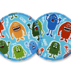 Könnte beinhalten: Zwei runde Stoffuntersetzer mit hellblauem Hintergrund und einem Muster aus bunten Cartoon-Monstern. Die Monster sind in verschiedenen Farben, darunter Orange, Grün und Blau, mit weißen Zähnen und mehreren Augen. Die Untersetzer haben einen blau genähten Rand.