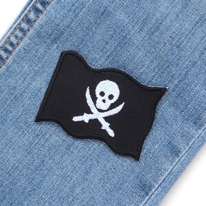 Peut inclure: Un &eacute;cusson de drapeau pirate noir avec un motif de cr&acirc;ne et d'os crois&eacute;s blancs cousu sur un tissu en jean bleu.