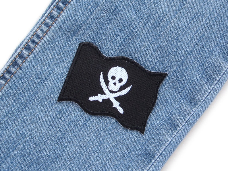 Peut inclure: Un &eacute;cusson de drapeau pirate noir avec un motif de cr&acirc;ne et d'os crois&eacute;s blancs cousu sur un tissu en jean bleu.