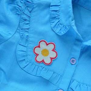 May include: A light blue button-up shirt with a white ruffle collar and a flower applique. The flower is white with a yellow center and a pink outline.