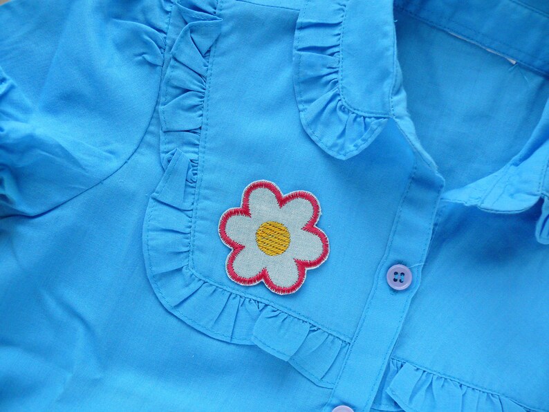 May include: A light blue button-up shirt with a white ruffle collar and a flower applique. The flower is white with a yellow center and a pink outline.