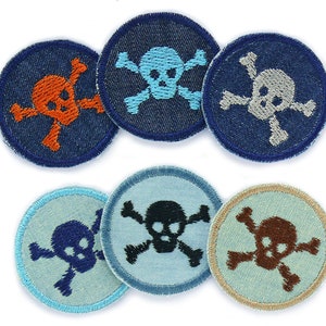 Op de afbeelding: Zes geborduurde strijkpatches met een schedel en gekruiste beenderen ontwerp. De patches zijn in verschillende kleuren, waaronder blauw, oranje, grijs, zwart, bruin en wit.