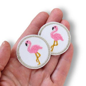 Peut inclure: Deux écussons brodés circulaires représentant un flamant rose avec un bec noir et des pattes jaunes sur un fond blanc. Chaque écusson possède une bordure cousue de couleur claire, créant un contour net et défini pour ces appliqués en tissu.