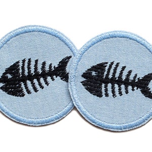 Può includere: Due toppe rotonde in tessuto blu chiaro con scheletri di pesce ricamati in nero.