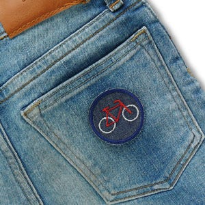 May include: Close-up of light blue denim jeans with a brown leather patch and a back pocket. The pocket features a round patch with a red bicycle design and white wheels, stitched on a navy blue background.