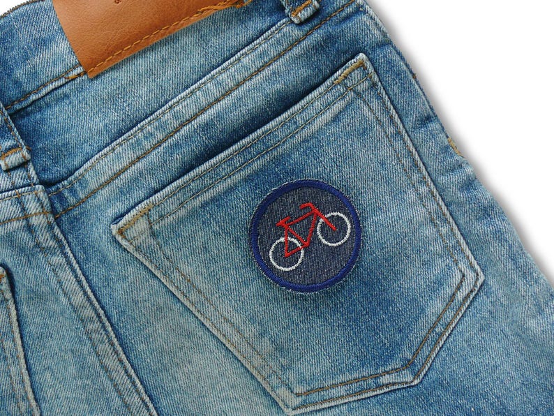 May include: Close-up of light blue denim jeans with a brown leather patch and a back pocket. The pocket features a round patch with a red bicycle design and white wheels, stitched on a navy blue background.