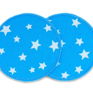 Peut inclure: Deux coussinets d'allaitement en tissu bleu avec des motifs d'étoiles blanches. Les coussinets sont ronds et ont une surface douce et texturée.