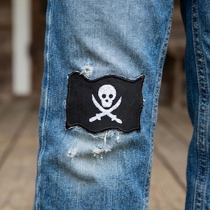 May include: A pair of light blue denim jeans featuring a rectangular black patch with a white embroidered skull and crossbones design. The patch is stitched onto the fabric, which shows signs of intentional distressing and fraying around the edges.