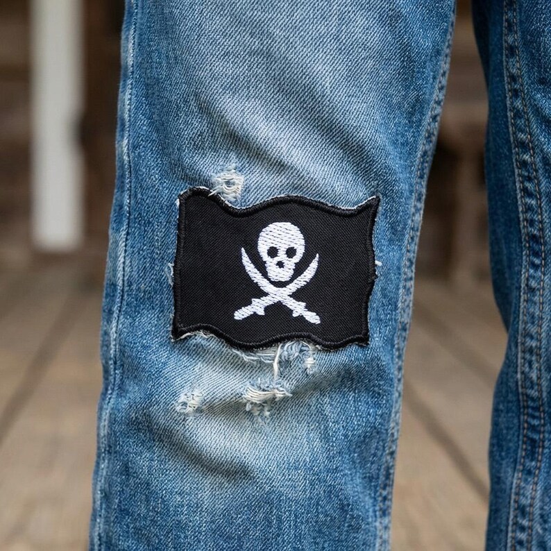May include: A pair of light blue denim jeans featuring a rectangular black patch with a white embroidered skull and crossbones design. The patch is stitched onto the fabric, which shows signs of intentional distressing and fraying around the edges.